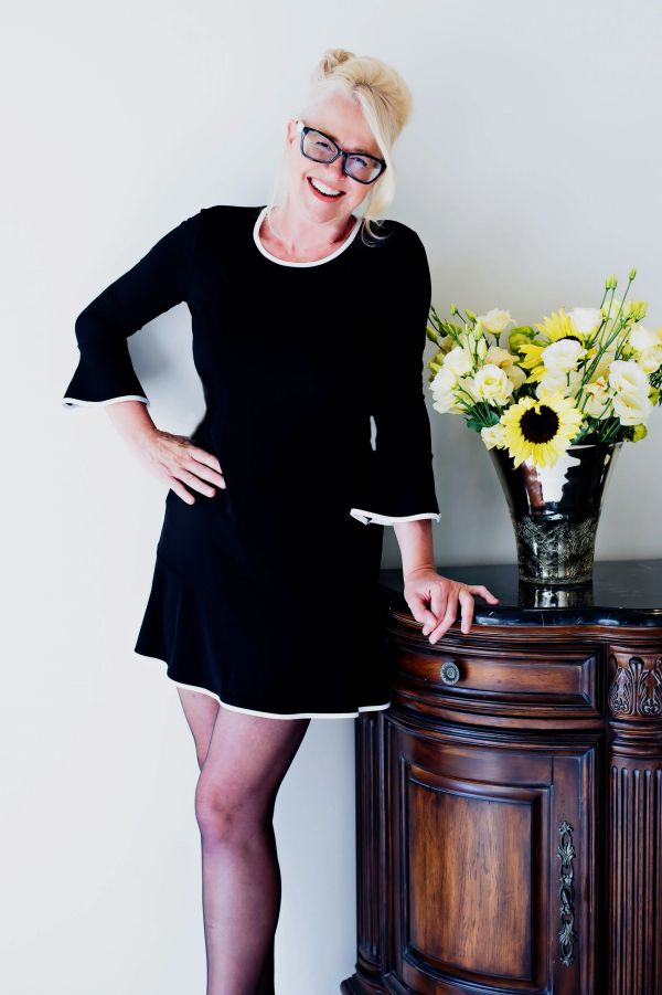 Smiling woman in black dress posing by a wooden cabinet with a vase of yellow flowers.