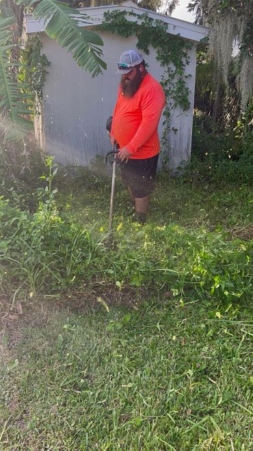 Team member using a string trimmer to clear dense vegetation near a fence.