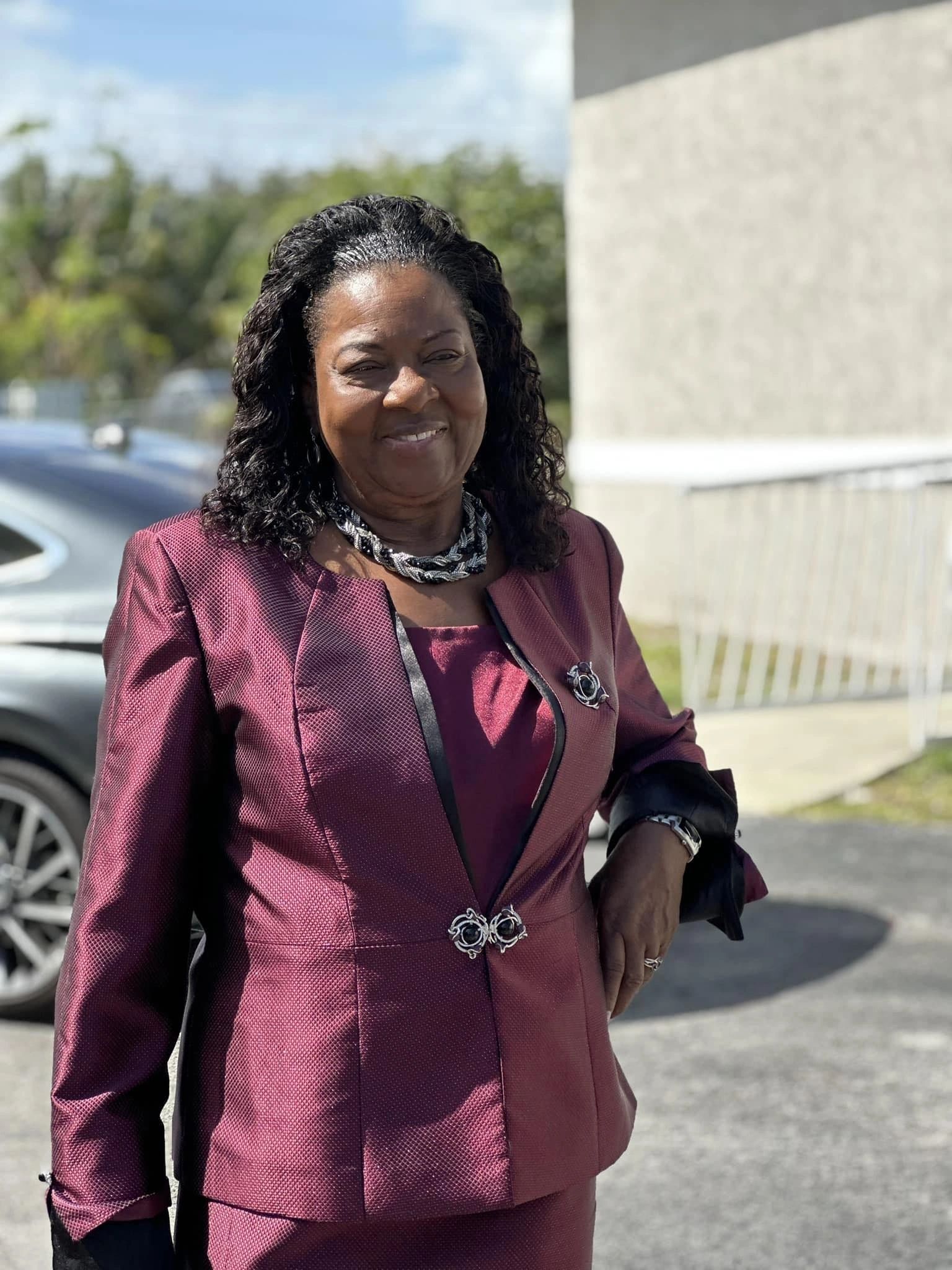 Woman in a maroon suit smiling outdoors on a sunny day.