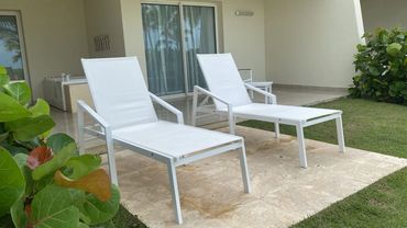 Two white lounge chairs on a patio with greenery around.