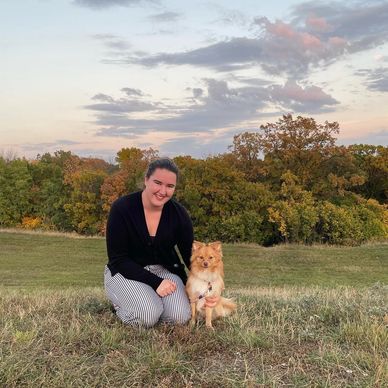 A woman kneeling on grass with her small dog during sunset.