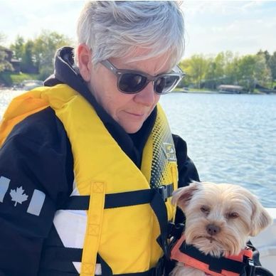 Person wearing sunglasses and a yellow life jacket holding a small dog on a boat.