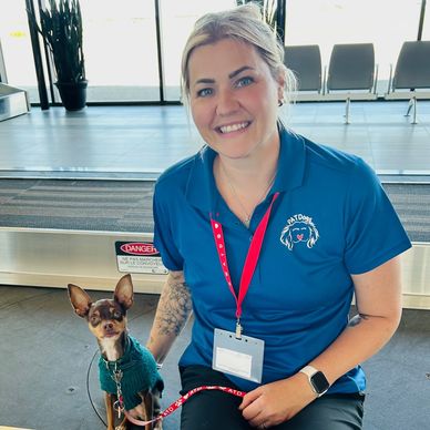 Woman in blue shirt sitting with a small dog in a green sweater at an airport.