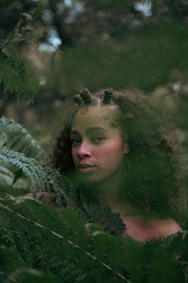 Woman with curly hair among lush green ferns, gazing thoughtfully.