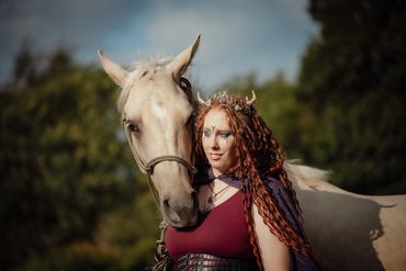 Woman with dreadlocks and fantasy makeup stands beside a white horse outdoors.