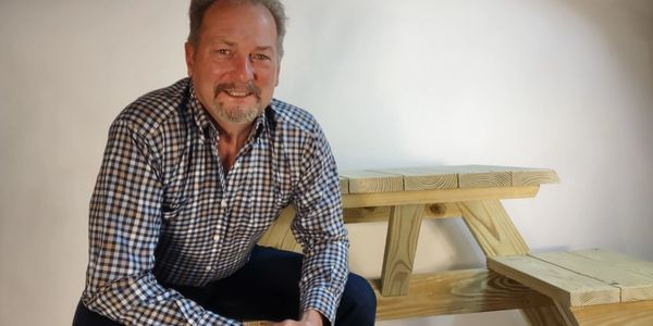 Smiling man in checkered shirt beside a wooden picnic table.