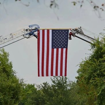 Large American flag suspended between two aerial ladders above trees.