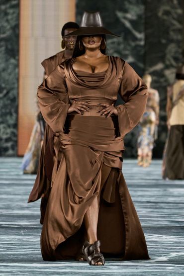 Model in a flowing brown satin dress with wide sleeves and a matching hat on the runway.