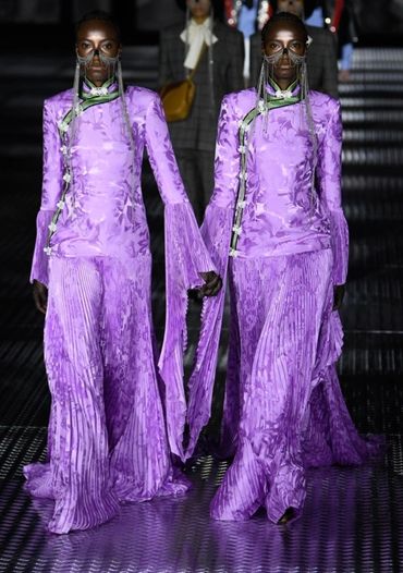 Two models in matching flowing purple outfits with face accessories on runway.
