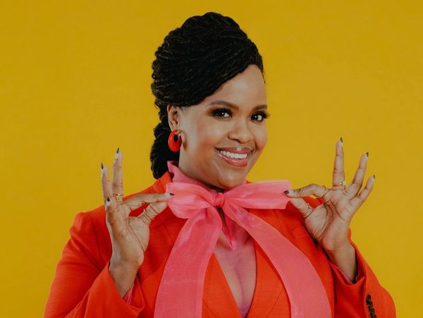 Smiling woman in a red outfit with a large pink bow against a yellow background.