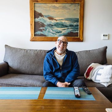 Smiling elderly man sitting on a couch with a seascape painting behind him.