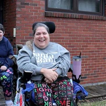 Two women sitting outside near a brick building, one in a wheelchair smiling.