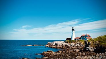Lighthouse on rocky coastline under blue sky and calm sea.