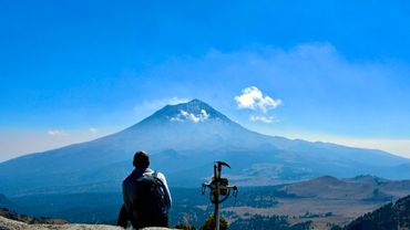 A person sits on a rock, admiring a majestic mountain under a clear blue sky.