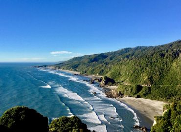 A scenic coastal view with waves crashing on a forested shoreline under a clear blue sky.