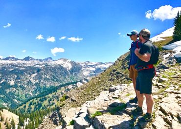 Two hikers stand on a rocky mountain trail overlooking snow-capped peaks.