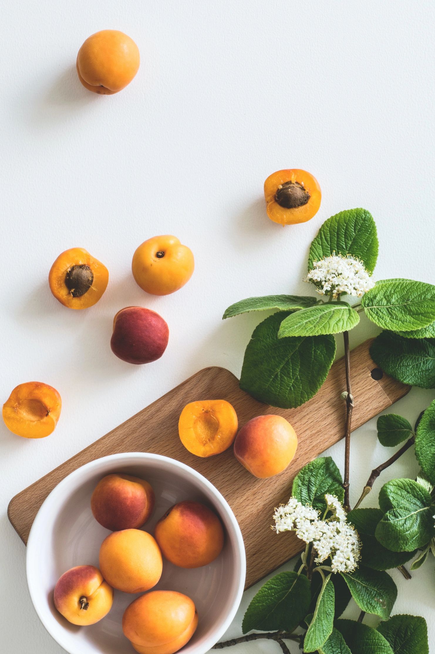 Apricots, some halved, scattered on a white surface with green leaves and flowers.