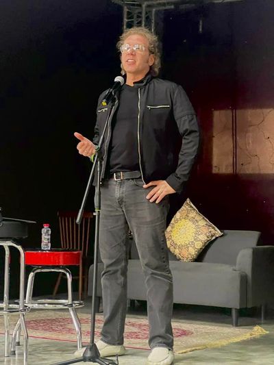 Man in black jacket speaking on stage with microphone, standing confidently.
