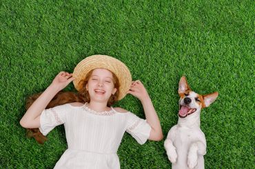 A joyful girl and her happy dog lie on green grass, both smiling and relaxed.