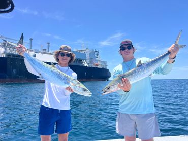 Two young men proudly holding large fish with a ship in the background.