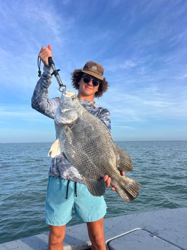 Young man proudly holding a large fish on a boat under a clear blue sky.