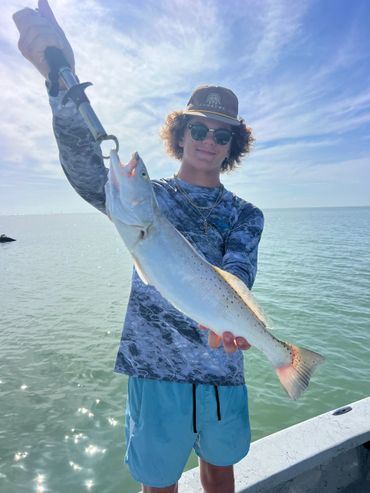Young man proudly holding a large fish on a boat under a bright blue sky.