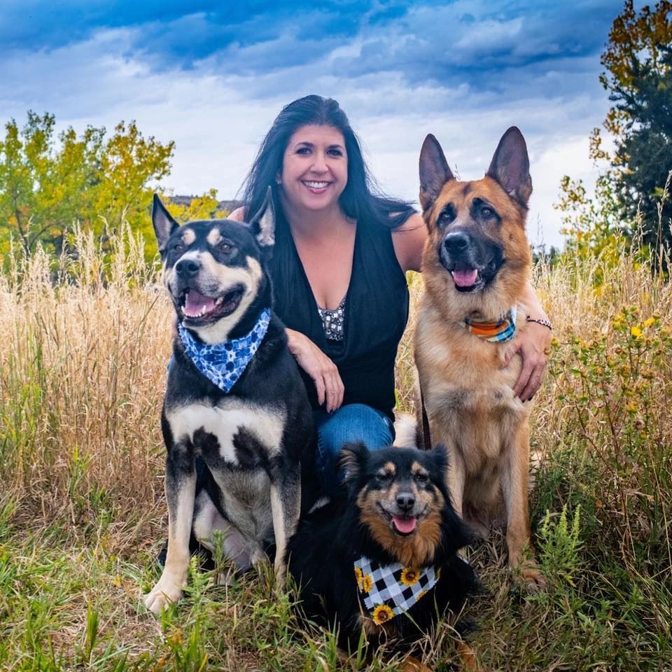 A smiling woman posing outdoors with three happy dogs wearing bandanas.