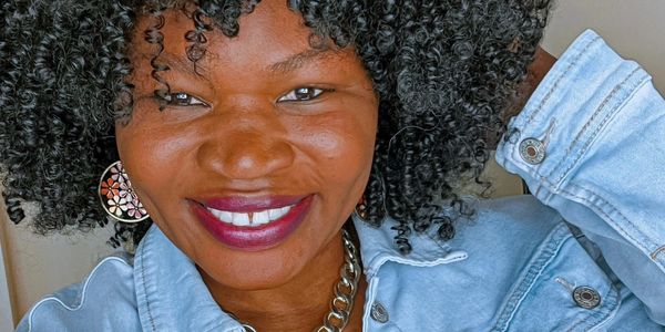 Woman with curly hair smiling, wearing denim jacket and chain necklace.