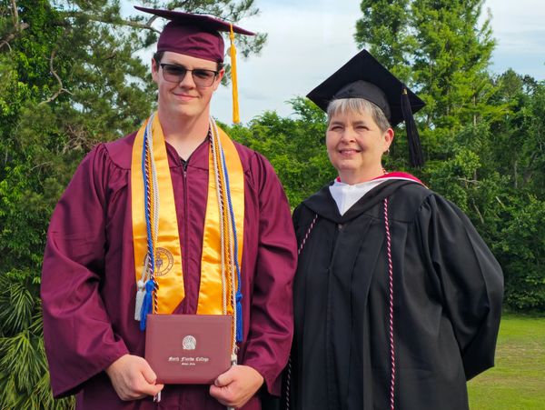 Graduate in maroon gown with diploma, beside a smiling faculty member in black academic regalia outdoors.