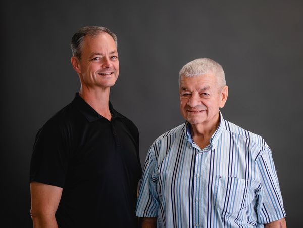 Two men smiling against a dark gray background.