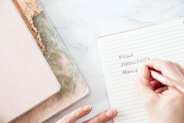 Notebooks and journals in a pile, with a woman's hand writing focus, productivity, family.