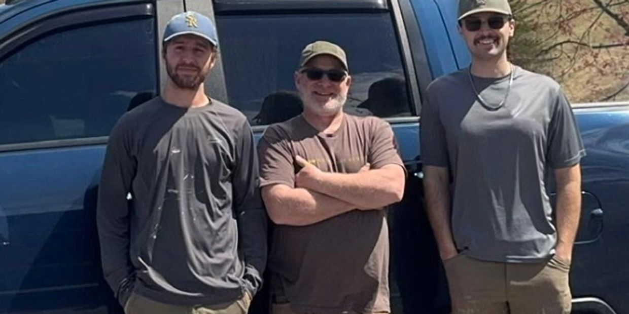 Three men posing in front of a blue vehicle, wearing casual outdoor attire and caps.