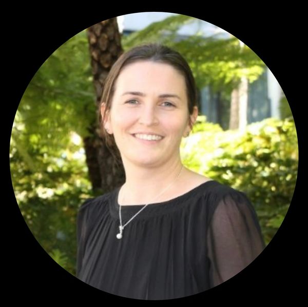 Smiling woman in a black blouse outdoors with greenery in the background.