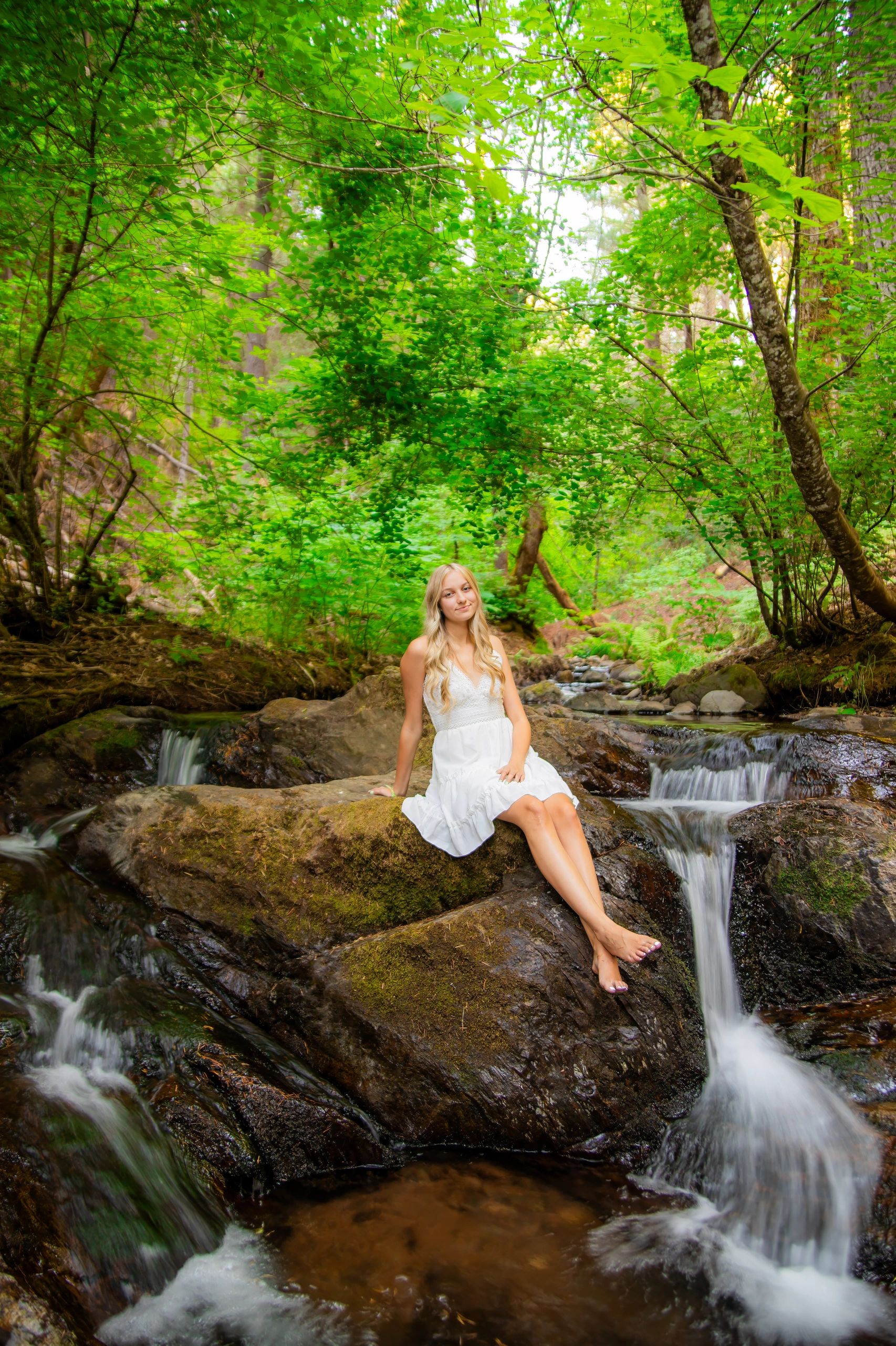 A young lady in a white dress relaxing on rocks amongst tranquil waterfalls, surrounded by dogwoods.