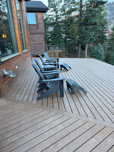 Four black Adirondack chairs with footrests on a wooden deck surrounded by pine trees.