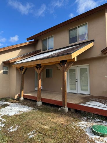 Wooden patio cover with light snow on roof and ground outside a beige house.