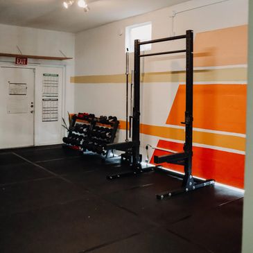 A gym corner with weight racks and a squat rack against an orange-striped wall.