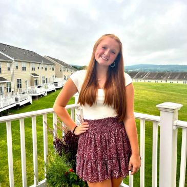 Smiling young woman posing on a porch with houses and greenery in the background.
