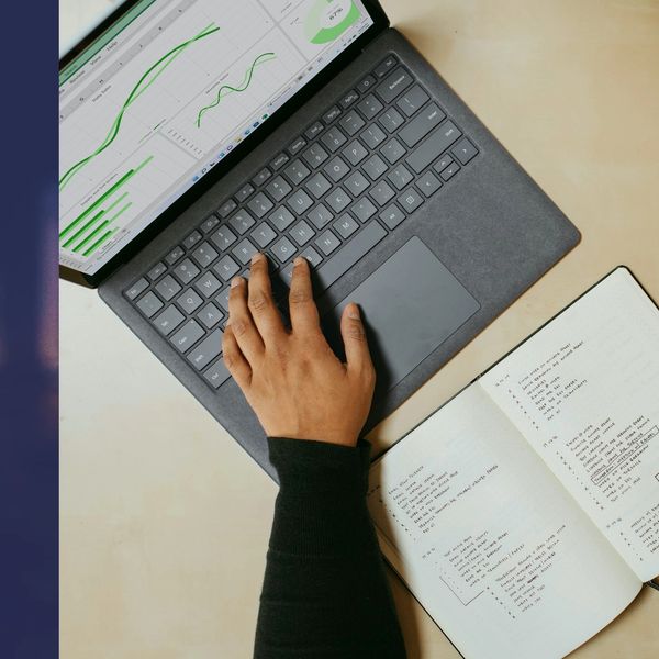 Laptop op tafel met de handen van een persoon. 1 hand op de laptop, de andere hand schrijft