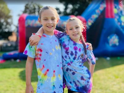 Two smiling kids in tie-dye shirts with arms around each other outdoors.