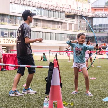 A young girl practices archery outdoors under adult supervision in an urban area.
