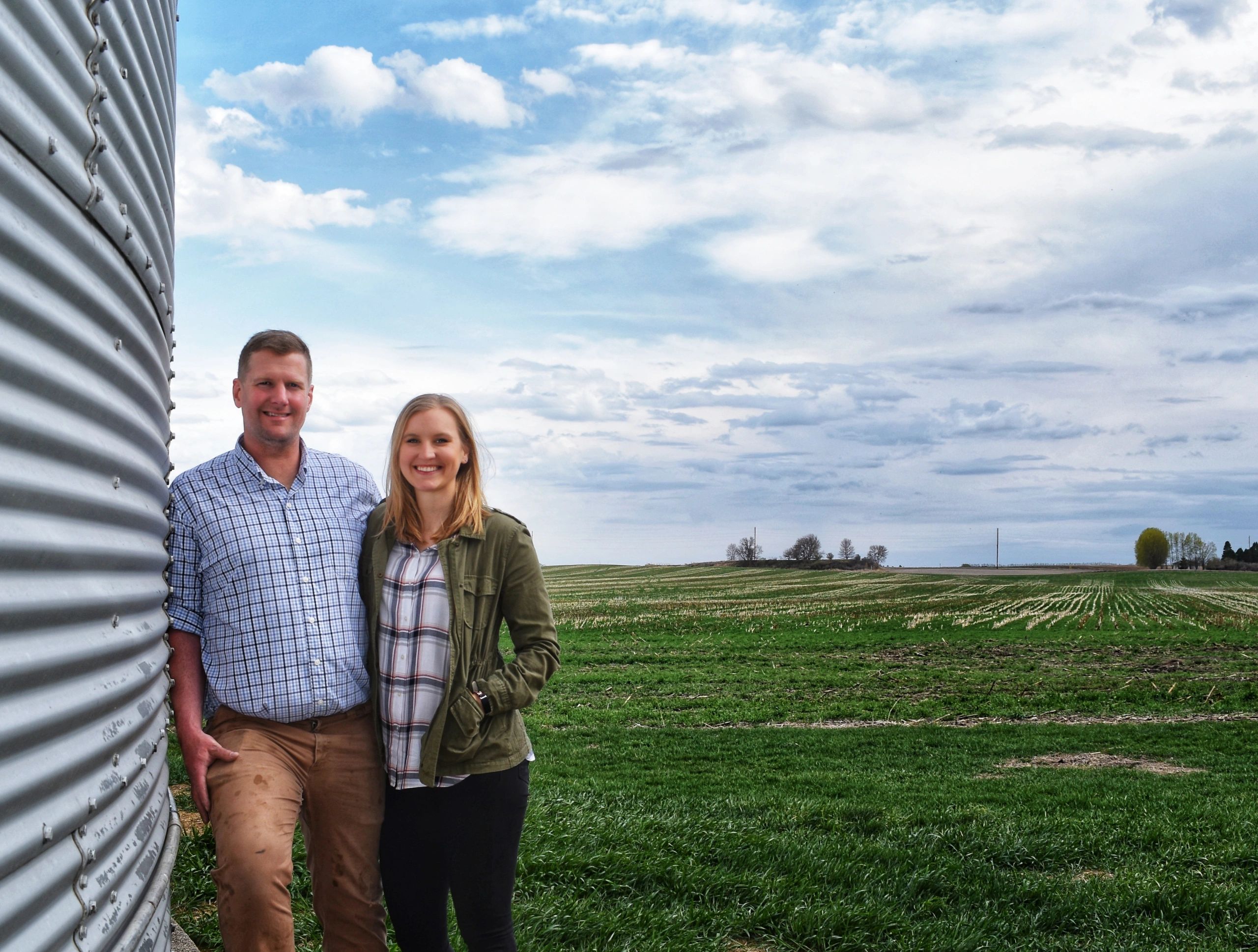 A smiling couple standing next to a grain silo in a rural field.