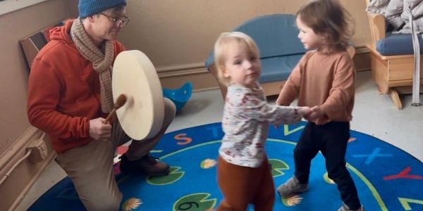 Adult playing drum while two toddlers dance on a colorful rug.
