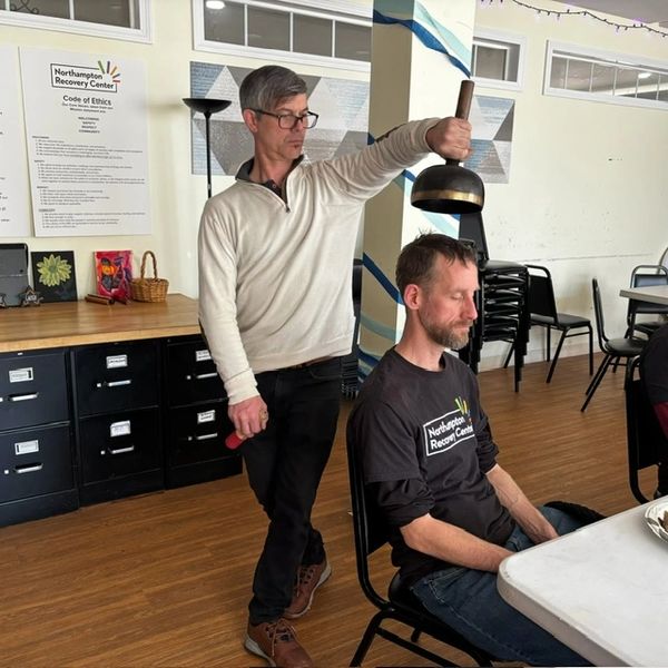 A man stands behind another man holding a large bell above his head in a community center.