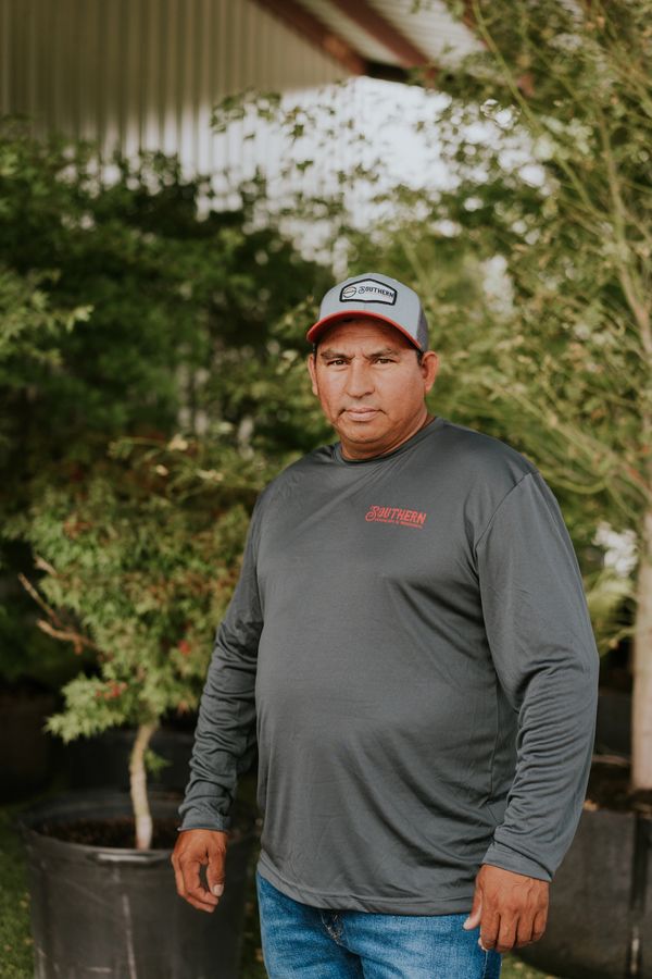 Man in a gray long-sleeve shirt and cap standing outdoors near plants.