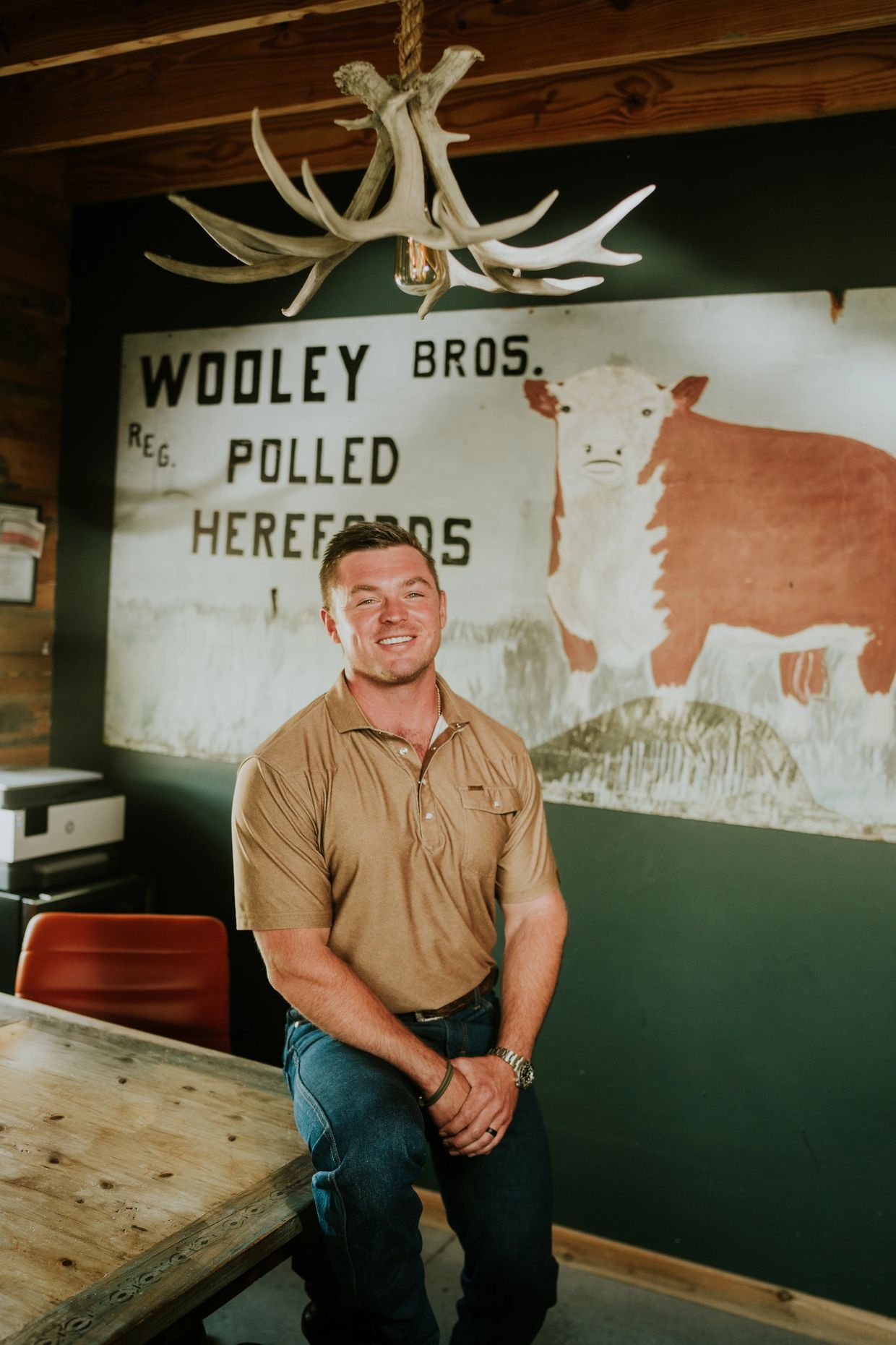 Smiling man sitting on a wooden table in a rustic room with cattle sign.
