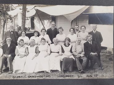 Portrait of 18 adults in front of an unknown structure. Curiously the man seated in the front row on