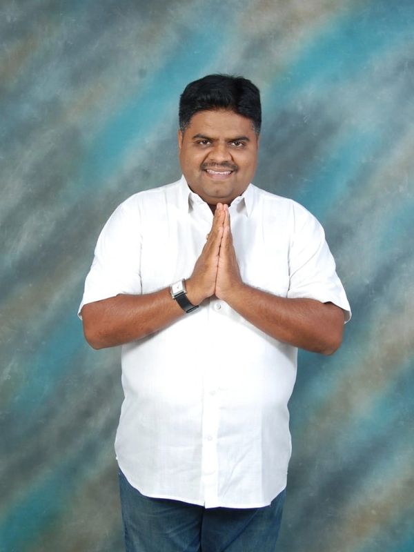 Man in white shirt greeting with folded hands against a blue-gray background.