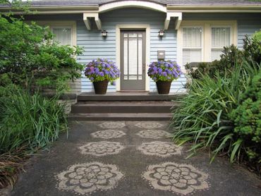 Front porch of a blue house with floral-patterned pavement and potted purple flowers.