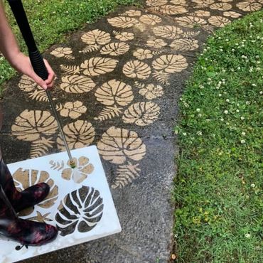 Person using stencil and water to create leaf patterns on a wet pathway.